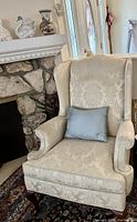 Front angled view of a clawfoot wingback chair upholstered in cream floral damask fabric with a blue throw pillow, next to a fireplace.