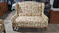 Front view of vintage floral love seat couch with wood legs, cream fabric with pink and green floral pattern.