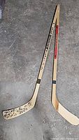 Two wooden hockey sticks laid on concrete ground. One stick has multiple black autographs on the blade and text along the shaft indicating Signature Series.