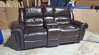 Front view of dark brown two-seater leather recliner couch showing plush cushions, armrest nailhead details, and center console with cup holders.