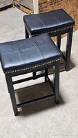Two black leather padded stools with wooden legs and silver stud detailing viewed diagonally from above showing seating surface and structure.