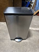 Front view of stainless steel rectangular trash can with foot pedal and closed lid, showing some surface scratches