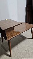 Side view showing the two-tier tabletop design with sculpted wooden supports and angled legs with caps at feet. Wood grain and brown finish visible.