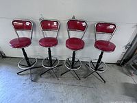 Set of four vintage chrome bar stools with red vinyl seats and backs, mounted on newer black metal bases with footrests, arranged in a row against a wall