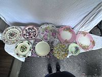 Overview of nine antique decorative plates arranged side by side, displaying various floral and vintage decorative patterns including two matching pink floral plates with scalloped edges.