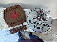 Two bar signs side by side on a white surface: a round Budweiser Beer sign and a wooden Molson Canada sign with hanging plaque.
