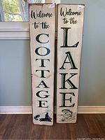 Front view of two wooden signs, "Welcome to the Cottage" and "Welcome to the Lake," standing side by side against a wall.