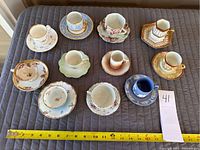 Top-down view of all 11 demitasse cups and saucers laid out, showing different styles and colors including blue, green, pink, and gold accented pieces.