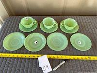 Three jadeite cups with handles aligned above three matching saucers and three small plates on a quilted surface, measuring tape for size reference.