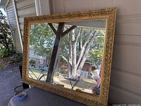 Full shot of ornate large gold framed mirror showing reflective glass and outdoor background.
