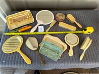 Lot of vintage French white vanity ware including trays, hand mirrors, hair brushes, and combs laid out on a quilted surface with measuring tape for scale.