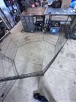 Top-down angled view showing the octagonal setup of 6 large black metal wire panels connected together forming the dog pen. Panels have a door visible.