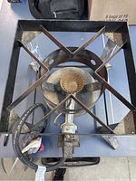 Top-down view of propane burner showing circular brass burner within square metal frame and connected rubber hose.