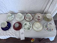 Photo showing 10 teacups with matching saucers arranged in two rows on a white cloth, various floral patterns and colors visible