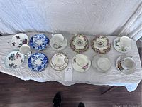 Overview of the 8 teacups and assorted mismatched saucers showing floral and oriental patterns on white cloth background.