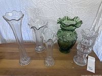 Five flower vases arranged on a wooden surface with a crumpled white fabric backdrop. Four are clear glass and one is green glass with embossed designs and ruffled rims.