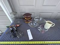 Photo showing all items in the lot laid out on a blue surface including two Pyrex glass bowls (one with measurement markings), two vintage Pyrex covered dishes with lids, glass canister with lid and copper scoop, two hand beaters, and a cream-colored ceramic pitcher.