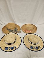 Four straw hats displayed on a white fabric background; two adult size hats with ribbons and two smaller child size hats with blue floral decorations on brim edges.