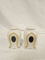 Front view of white ceramic salt and pepper shakers decorated with gold arches and crystal rhinestones, showing medallion detail on front.
