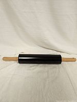 Photo of a lightweight rolling pin with a black metal-like cylindrical center and wooden handles on both ends, placed horizontally against a white textured fabric backdrop.