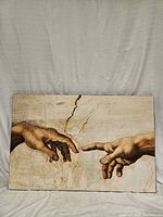 Front view of framed artwork showing close-up of two hands from Michaelangelo's Creation of Adam, with finger tips almost touching on a cracked beige background.