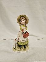 Front view of porcelain figurine showing girl in green dress with bonnet holding red basket and lamb at her side.