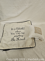 Photo of a cream colored 16x16 pillow cover with black piping featuring the quote 'In a World where You can be anything Be Kind' and a smaller 8x8 cream colored pillow with the text 'Love Note'.