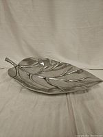 Silver metallic tray shaped like a leaf, showing full length and curved edges with stem, stood on small feet.