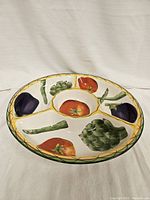 Top view of round ceramic serving tray with four compartments painted with colorful vegetable designs and green border.