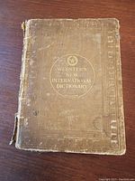 Front cover of the 1927 Webster's New International Dictionary showing wear, frayed fabric, detached cover and embossed gold lettering.