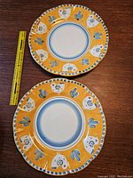 Two orange Solimene Vietri Campagna terracotta dinner plates with white roosters and blue floral motifs on orange borders, 12 inches size with measuring tape.