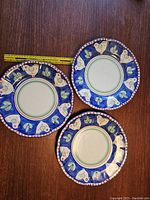Three blue Solimen terracotta dinner plates with white bird and green leaf decoration, positioned on wooden surface with measuring tape showing 12-inch size