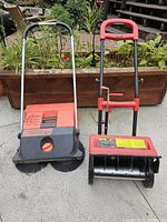 Haaga leaf sweeper and Spectra electric snowblower side by side outdoors on concrete with plants in background.