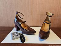 Front and side view of brown patent leather Fluevog heels with ankle straps, black plastic shoehorn and playing card box with John Fluevog image.