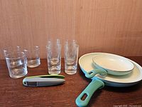 Overview of two frying pans, garlic press, and sets of glasses on wooden table.
