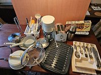 Overview of assorted kitchenware and small appliances on table including coffee maker, toaster, thermos, frying pans, grilling pan, metal fruit basket, kitchen utensils in caddy, and cutlery tray.