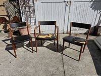 Three chairs grouped: one armchair at center flanked by two side chairs. All show torn black vinyl upholstery and exposed yellow foam.
