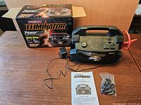 Photo showing MotoMaster Eliminator PowerBox power inverter with original box, charger cable, accessories, and owner's manual on table.