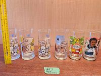 Photo showing six clear movie-themed drinking glasses with various colorful cartoon and character designs, all approximately 6 inches tall, with a measuring ruler for size reference and a lot number on green paper.