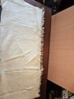 Photo showing the blanket unfolded partly on a dark wood surface, highlighting its off white wool texture and fringed edge.