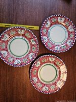 Three round terracotta dinner plates decorated with red birds, leaves and floral patterns with a blue dotted outer rim, shown with a measuring tape indicating approximately 12 inches diameter.
