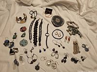 Overview of all jewelry pieces laid out on cloth including bracelets, necklaces, numerous pairs of earrings, and stone pendants.