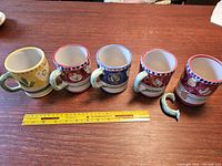 Five Vietri Solimene Campagna terracotta coffee mugs lined up on a wooden surface, showing colors and patterns. One handle is broken and placed beside the mugs.