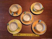 Top down view of 5 Vietri Solimene Campagna tea cups placed on matching saucers in yellow and orange with green and blue detailing around rims and handles, arranged in a circular pattern with measuring tape nearby for scale.