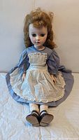 Full view of the vintage 1950s hard plastic doll wearing a blue dress with a cream checkered apron and curly brown hair.