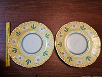 Two yellow terracotta dinner plates with hand-painted white birds, green leaves, and orange checkered rim design lying flat on a wooden surface next to a ruler showing the plate diameter.