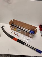 Saw blade with handle laid out next to disassembled pole sections in box, along with small hardware bag and instruction sheet.