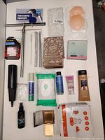 Top view of assorted household items arranged on a table including telescoping squeegee, straws, grout pen, personal care lotions, wipes, mirror and cosmetic sponges.