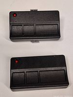 Two black programmable garage door remotes with three buttons and red LED indicator, front view showing flat button layout.