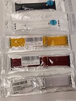 Five Apple iWatch bands and straps in original packaging, displaying black and white silicone rubber bands on top, yellow, red, and light gray nylon fabric bands below